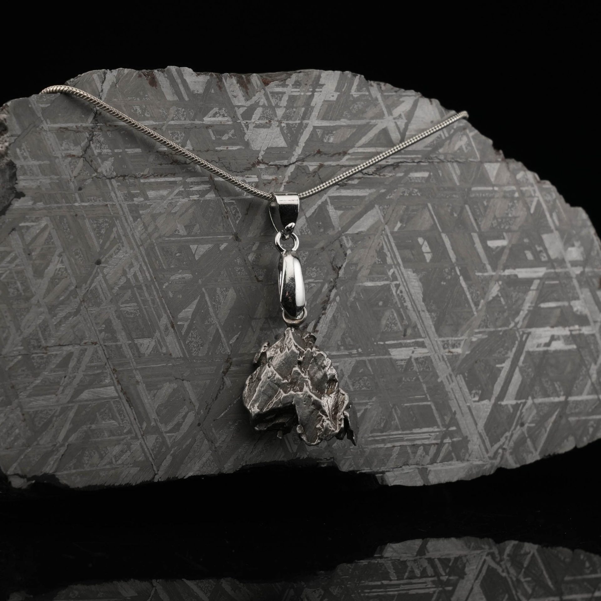 A silver meteorite necklace hangs on a large, patterned meteorite slice, with a black background and reflection.