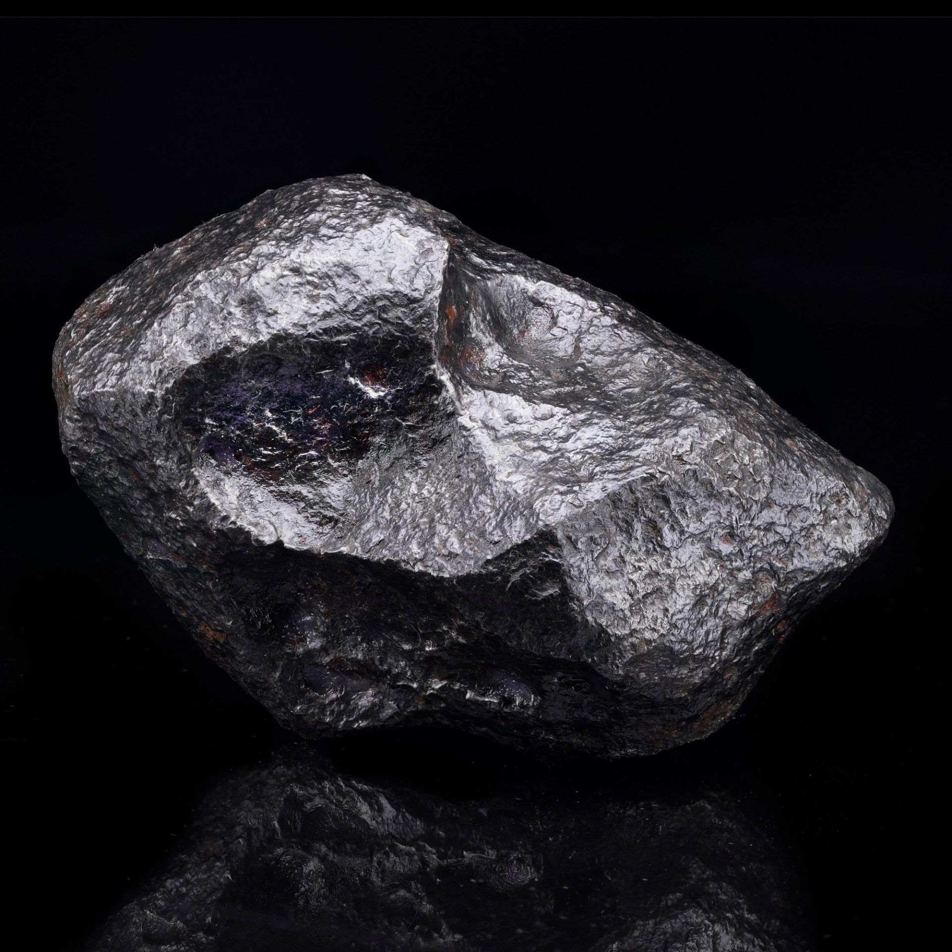 A close-up shot of a dark, metallic meteorite with a rough, textured surface and some colorful reflections.
