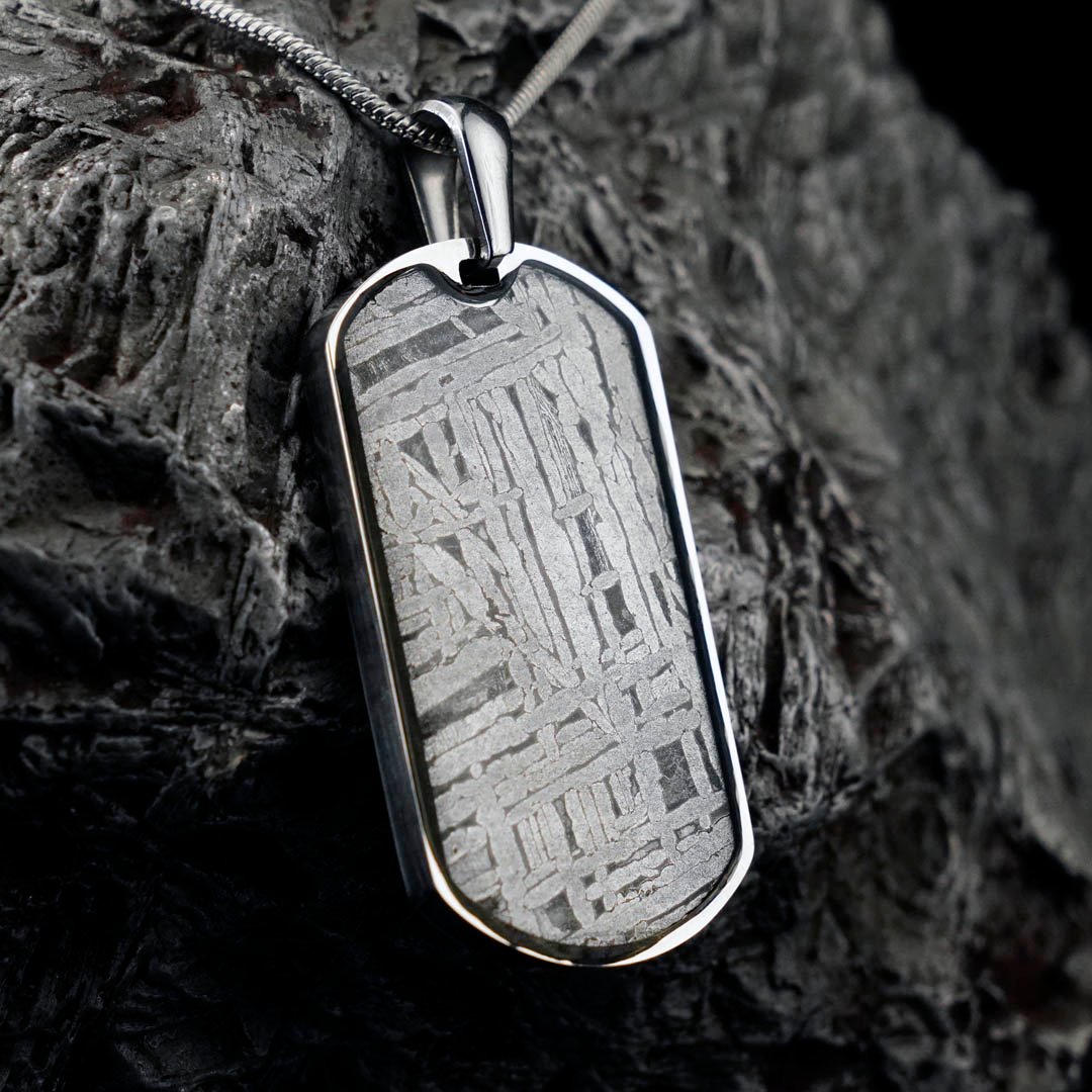 A silver dog tag pendant featuring a striking meteorite pattern rests on a textured dark rock.