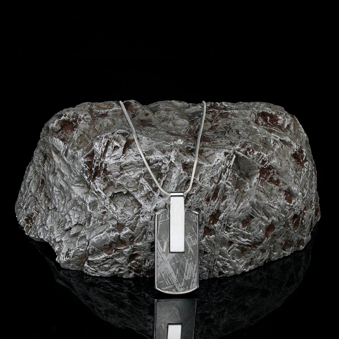 A silver necklace with a rectangular meteorite pendant rests on a large, textured meteorite rock against a black background.