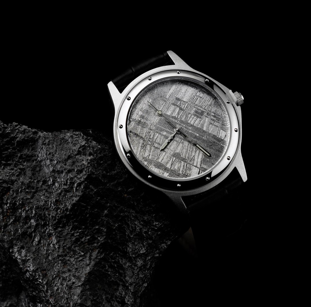 A close-up of a luxury watch with a patterned meteorite dial and a black strap, resting on a dark rock.
