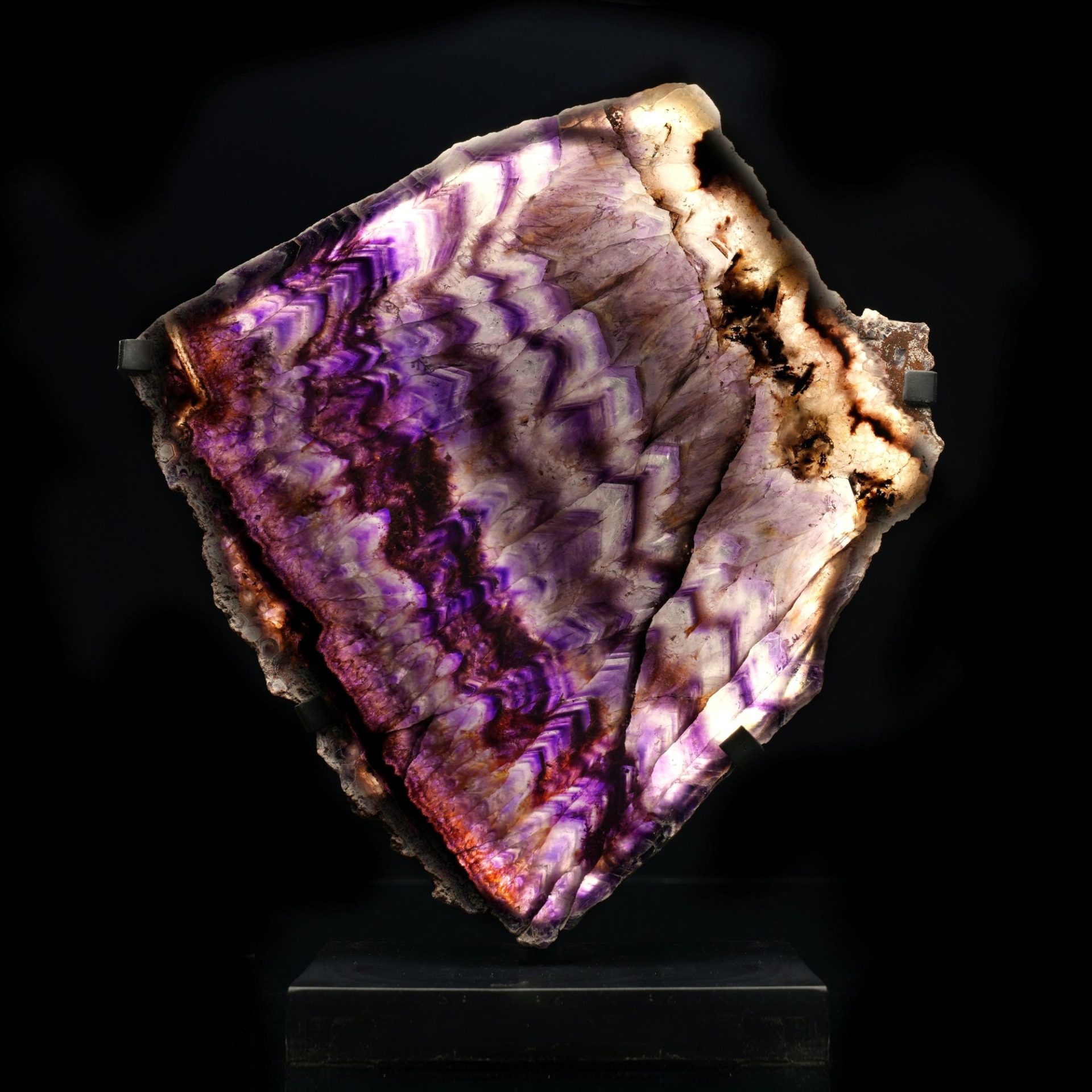 A stunning backlit amethyst crystal slice on a dark stand, showcasing vibrant purple and white chevron patterns.