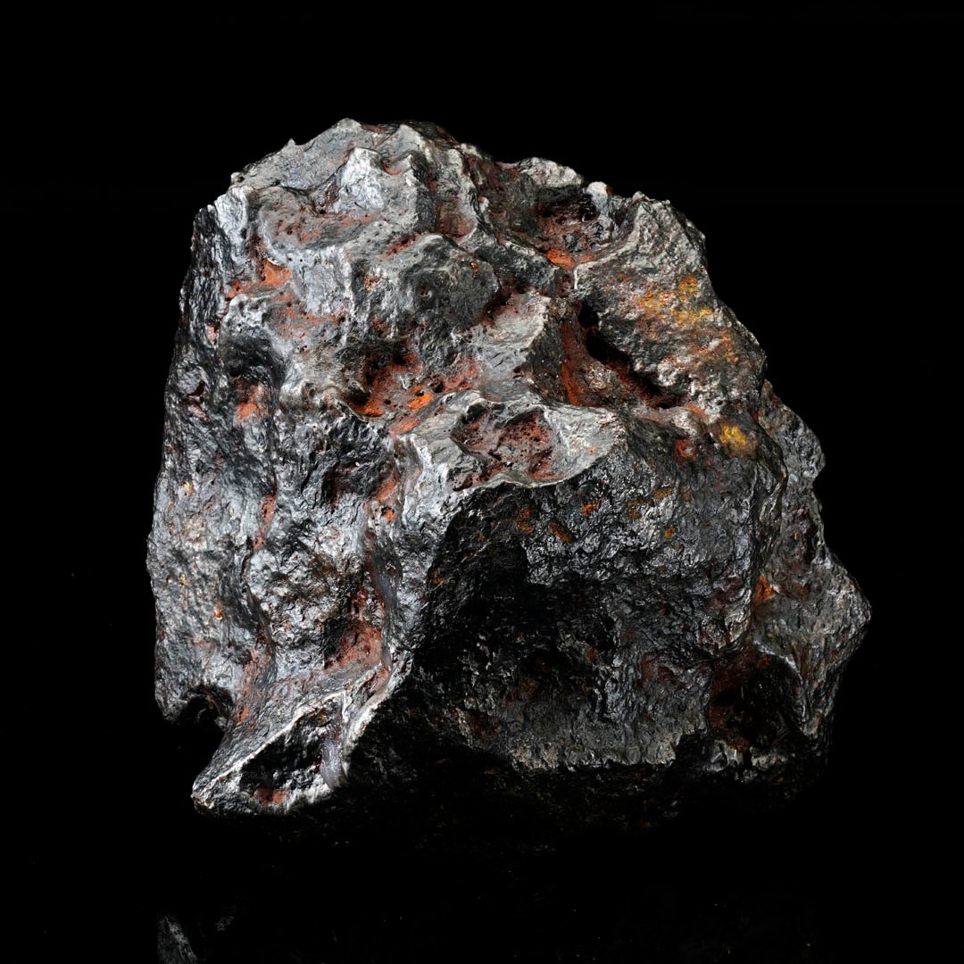 A dark, metallic, irregularly shaped meteorite with reddish-orange patches against a black background.