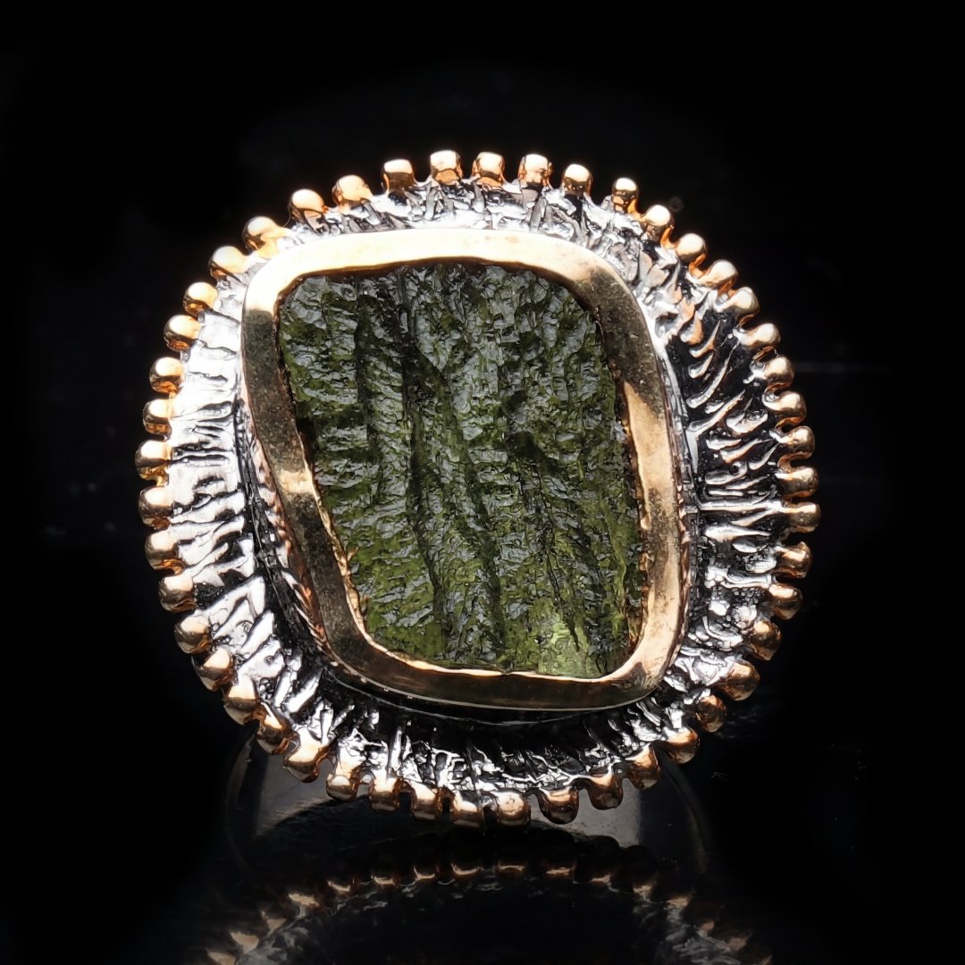 A detailed close-up of an ornate ring featuring a textured, rectangular green moldavite stone set in silver and gold-toned metal.