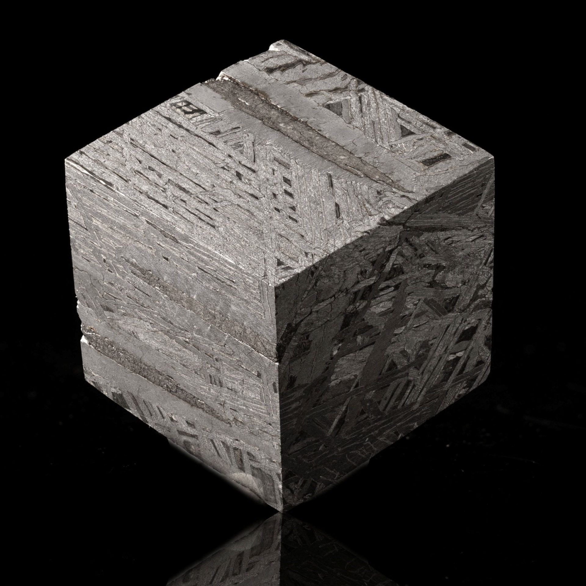 A polished, cuboid slice of a meteorite displaying intricate metallic Widmanstätten patterns on its surfaces, reflecting light.