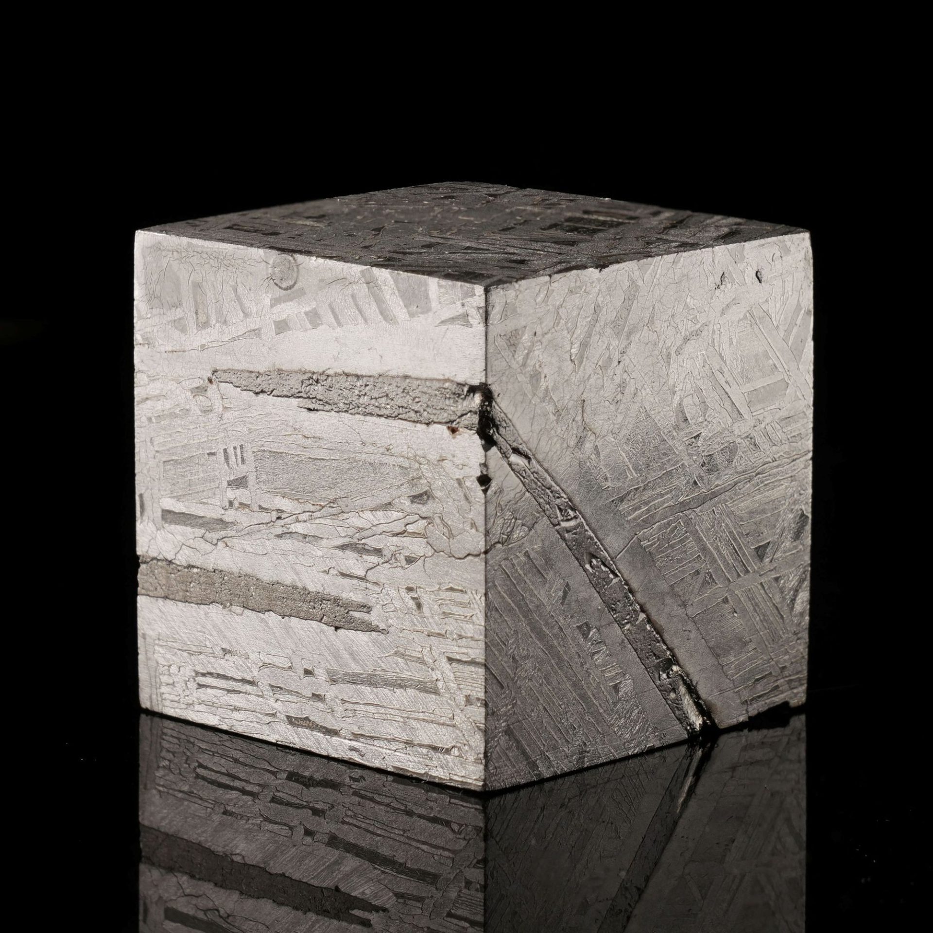 A cube-shaped meteorite with intricate metallic patterns on a dark, reflective surface.